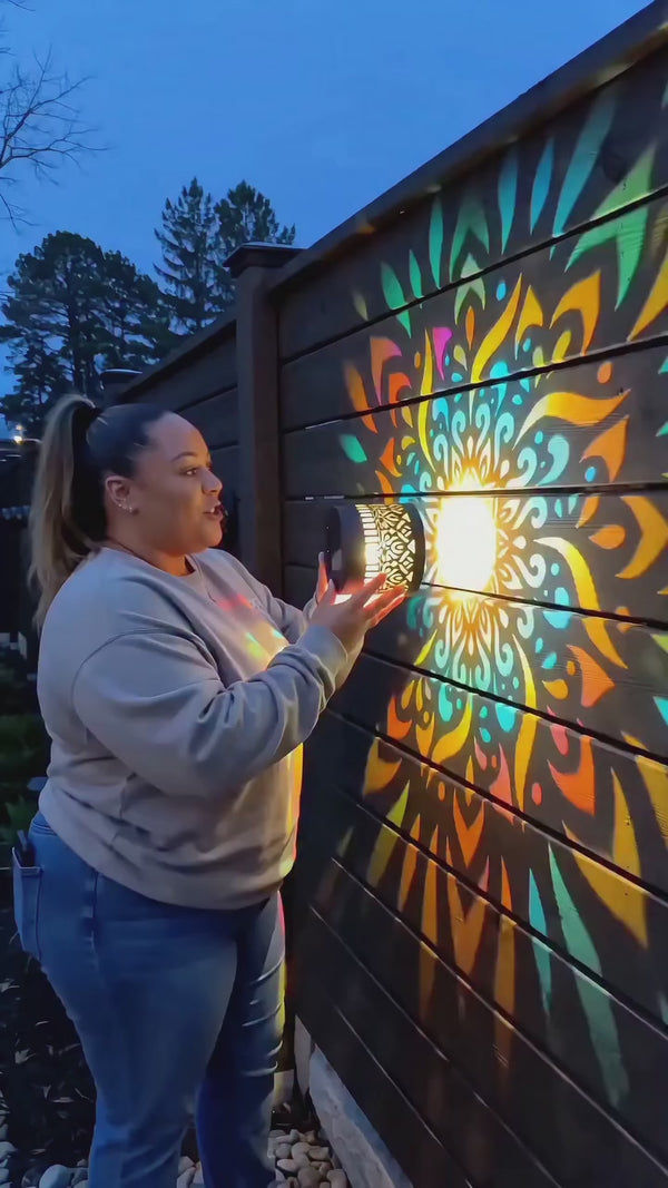 Woman holding a solar mandala lamp in front of a colorful projection on a wooden fence